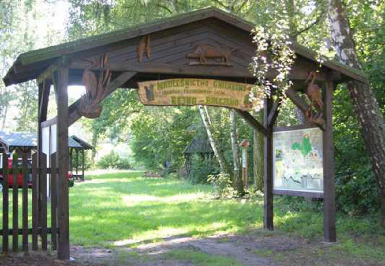 Drewniana brama w ciemnobrązowym kolrze z rzeźbami zwierząt, po prawej tablica z mapą, w tle zielona roślinność.