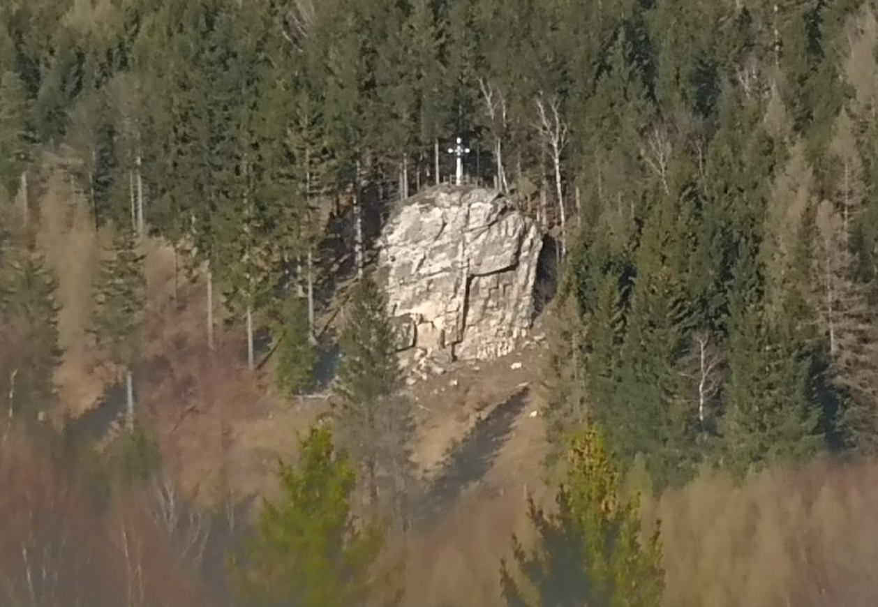 Ze wzniesienia porośniętego lasem wyłania się biała skała. Na niej krzyż. 